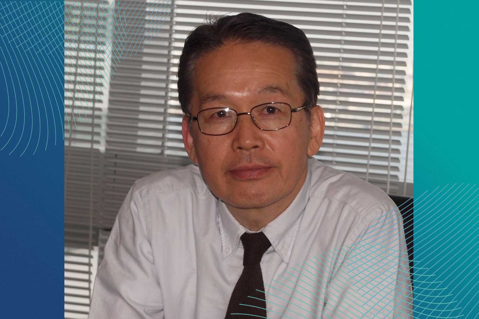 A person wearing a white shirt and a dark tie sits at a desk with blinds in the background.