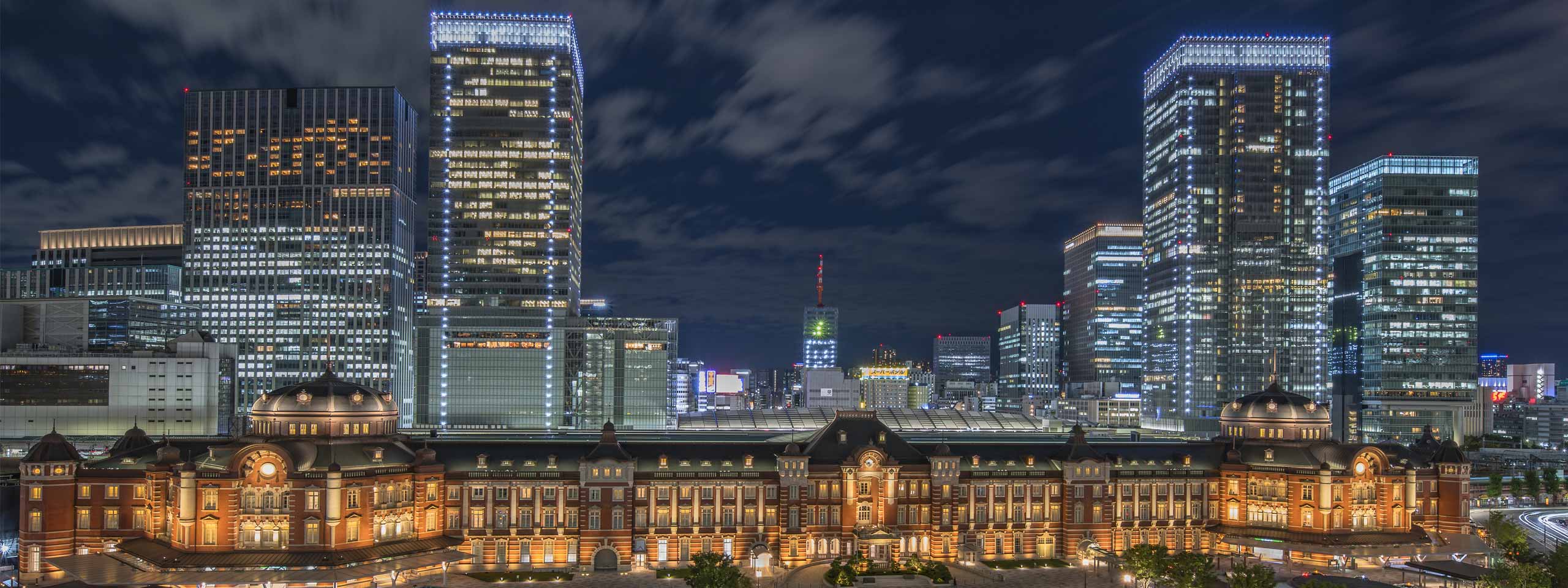 Tokyo Station illuminated at night, surrounded by modern skyscrapers and clouds in the evening sky.