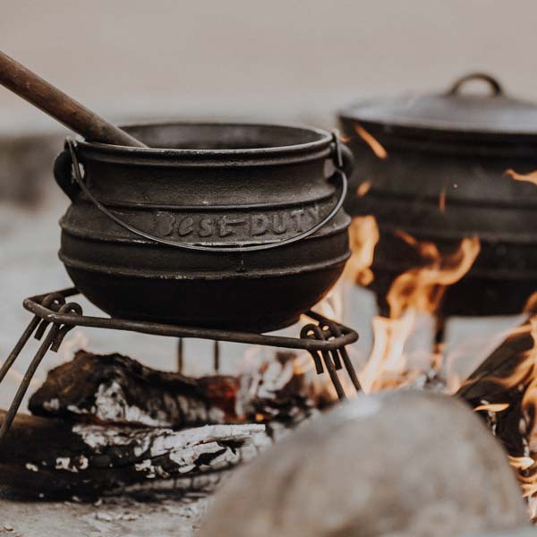 A black cast iron pot over a campfire, with a wooden ladle, surrounded by flames and logs.