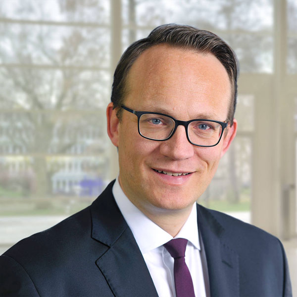 A man in a suit and tie stands in a modern, bright room with windows in the background, portrait of Dr Markus Krebber.