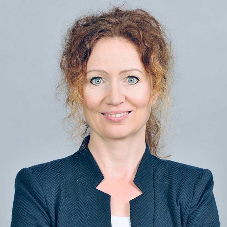 A person with curly red hair wears a black textured jacket and has their arms crossed. Portrait of Vera Bücker.
