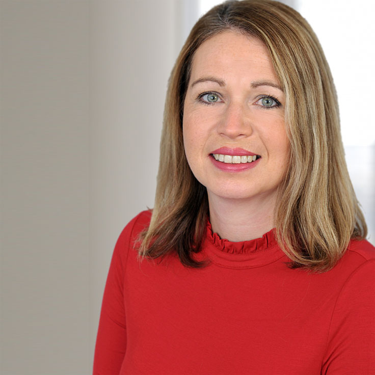 A person with long, straight hair wears a red top. The background is bright and softly focused. Portrait of Kelly Nye.
