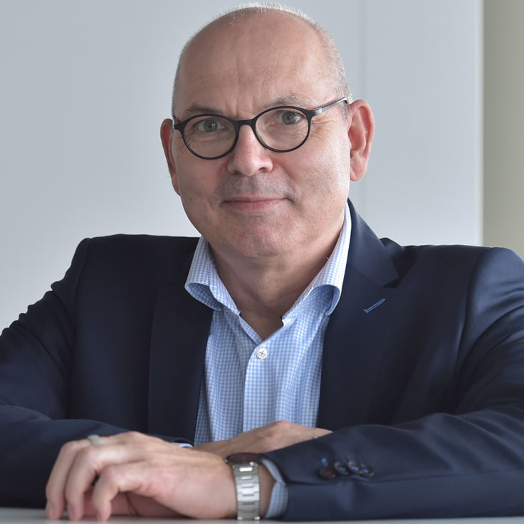 A man sits at a table, wearing a dark blue suit and a checked shirt. His hands are resting on the table. Portrait of Lothar Lambertz.